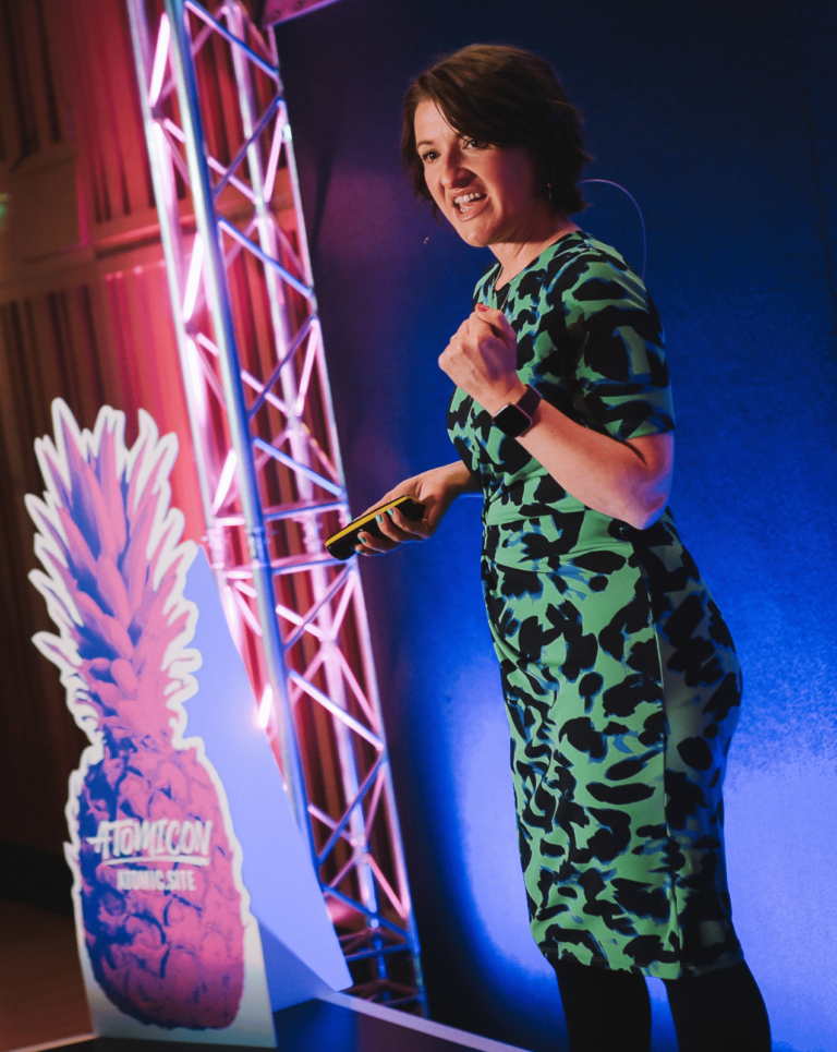 Janine on stage at small business marketing conference Atomoncon21 wearing a black and green dress. There are pink cardboard pineapples in the background with 'Atomicon' written on them.