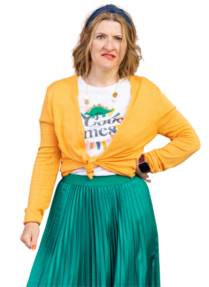 She wears a yellowy orange cardigan over a white slogan t-shirt and a bright green skirt. She is a middle aged woman with mid length, mid blond hair. She stands with one hand on her hip and looks concerned.