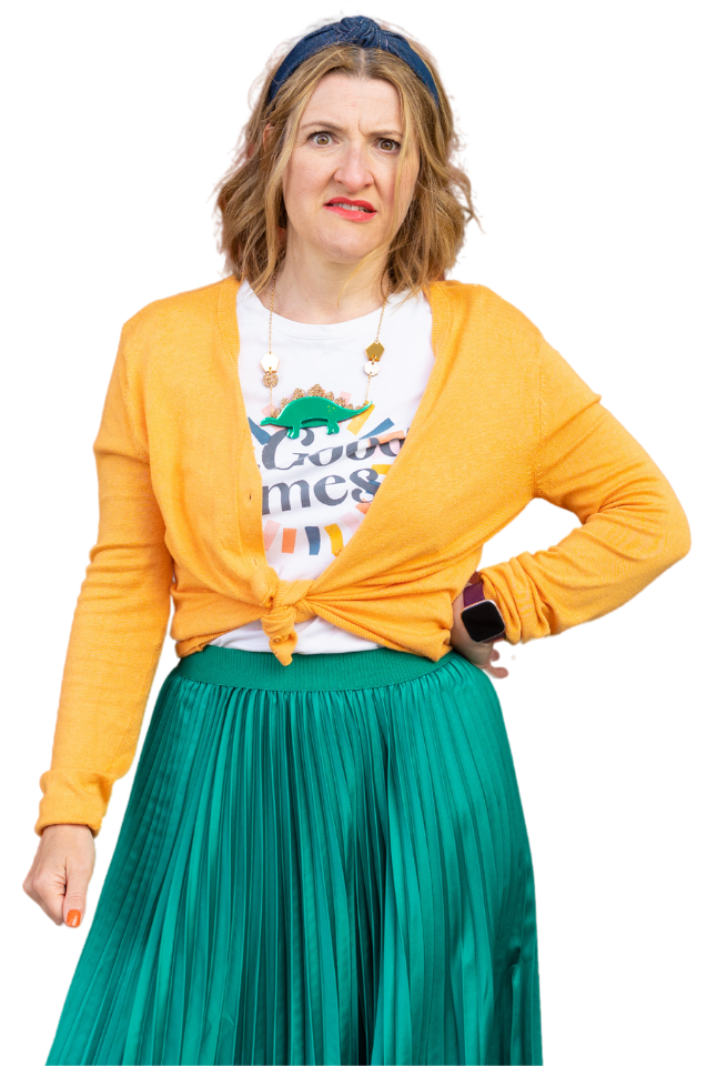 She wears a yellowy orange cardigan over a white slogan t-shirt and a bright green skirt. She is a middle aged woman with mid length, mid blond hair. She stands with one hand on her hip and looks concerned.