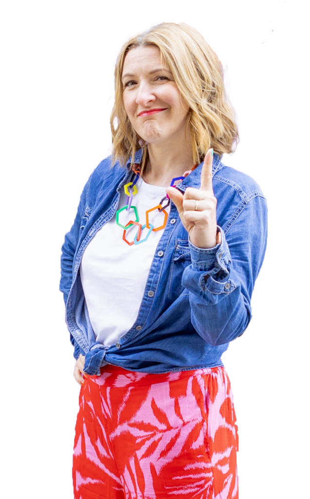 Janine Coombes stands three quarters on to the camera with one hand on her hib and the other with one finger pointing up like she’s about to make a point. She smiles with a knowing look. Janine is a middle-aged white woman with shoulder length mid-blonde hair she wears a denim shirt, red and pink summery trousers and white trainers.