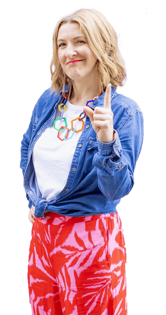 Janine Coombes stands three quarters on to the camera with one hand on her hib and the other with one finger pointing up like she’s about to make a point. She smiles with a knowing look. Janine is a middle-aged white woman with shoulder length mid-blonde hair she wears a denim shirt, red and pink summery trousers and white trainers.