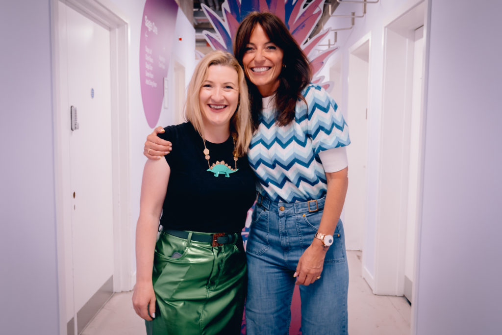 Janine Coombes stands to the left of the shot, Davina McCall on the right. Davina has her arm around Janine and her head is resting on Janine’s head. They’re both smiling broadly. Janine wears a shiny green pencil skirt, black t-shirt and stegosaurus necklace. Davina wears a pair of gorgeous high waisted flared jeans and a crocheted blue and white top. Both are middle aged white women. Janine has shoulder length mid-blonde hair. Davina has long brown hair. They’re in a corridor backstage at the Atomicon24 conference.