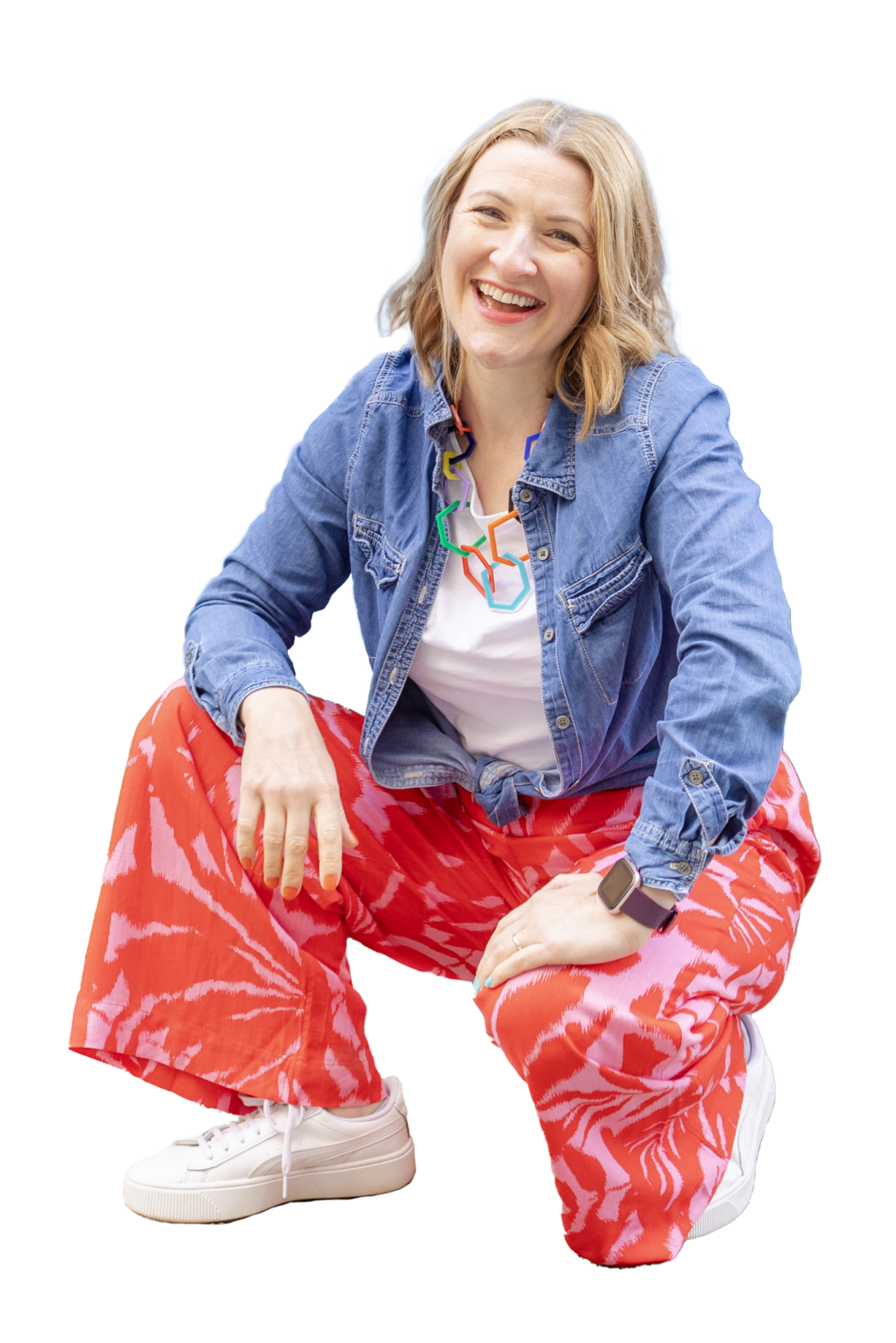 Janine crouches down with a hand resting on one knee. She is smiling at the camera. She's a white woman with shoulder length mid-blonde hair. She wears a denim shirt over a white t-shirt. Bright red and pink lightweight trousers and white trainers.