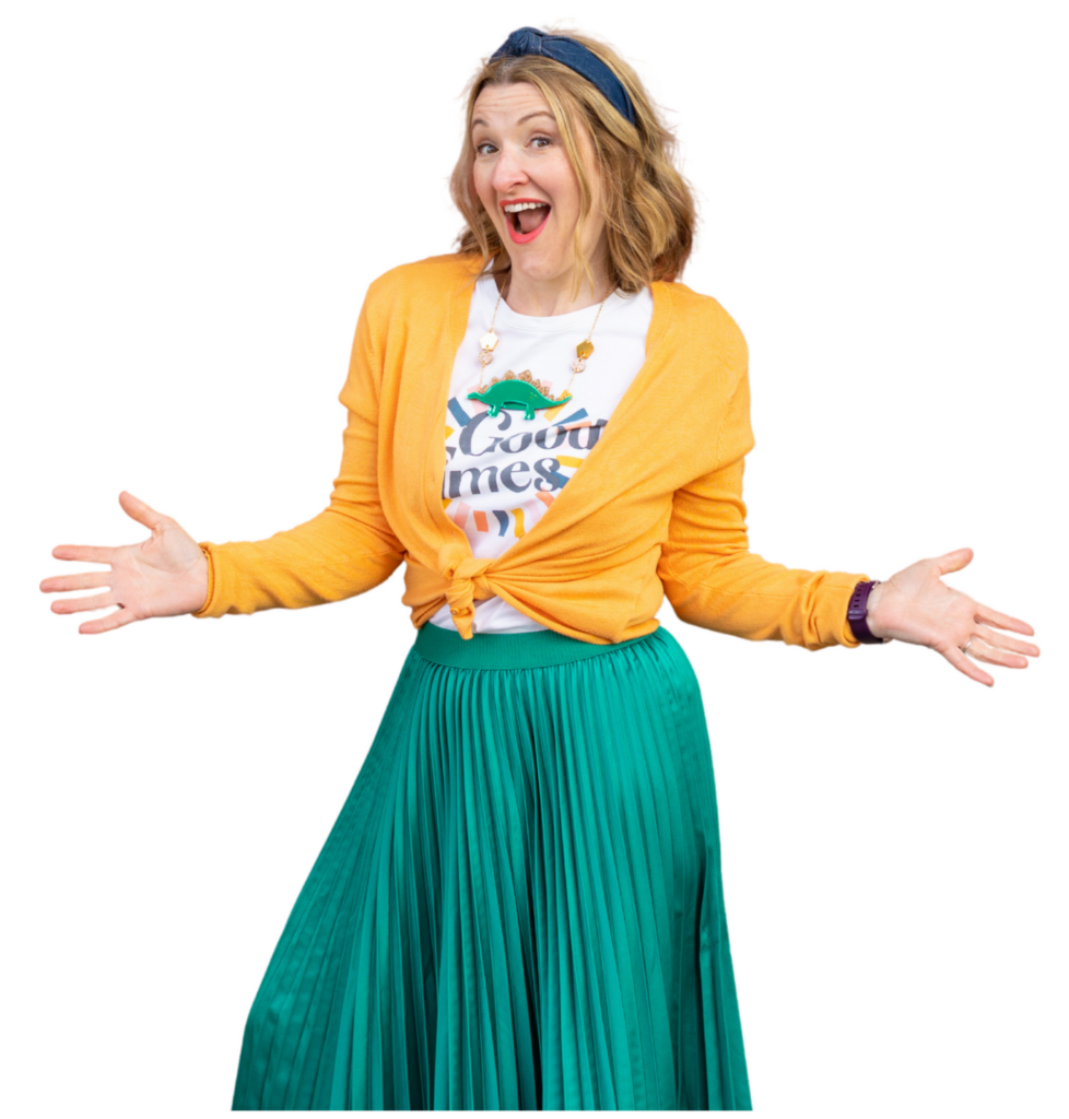 Janine Coombes, a middle-aged white woman with shoulder length dark blonde hair stands to the left with her hands out as if to say ‘ta dah!’ She wears a white t-shirt with ‘good times’ on it, a green pleated skirt and a yellowy orange cardigan.